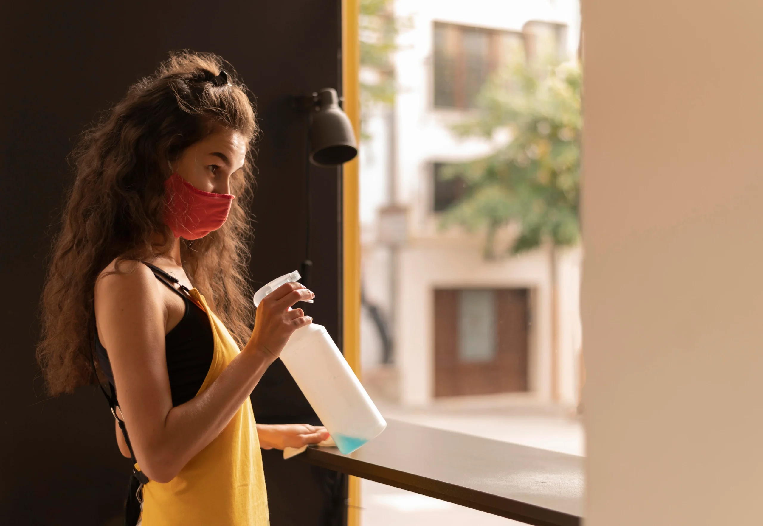 A woman wearing a mask holds a bottle, standing in a neutral background, conveying a sense of caution and health awareness.